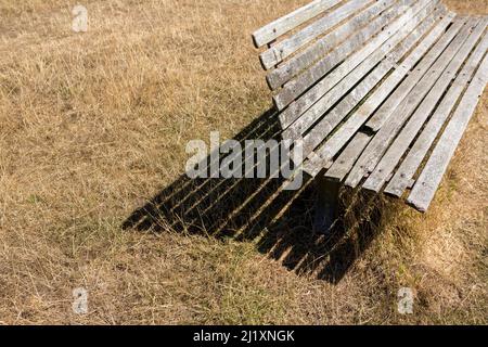 Una vecchia panca di legno in un po' di erba secca, sbiadita e morta, sbiancata quasi bianca sul caldo sole estivo. Foto Stock