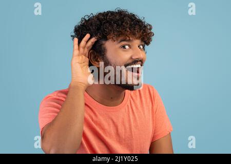 Curioso ragazzo indiano che tiene la mano per l'orecchio su blu Foto Stock