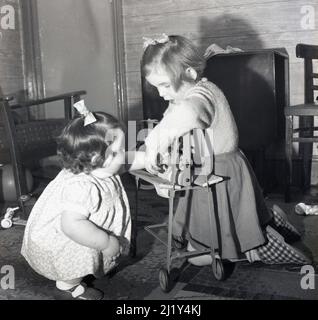 1950s, storica, una bambina e il suo piccolo assitore che gioca all'interno di una stanza con un giocattolo mangle, Inghilterra, Regno Unito. Il mangle giocattolo con telaio in metallo, con rulli in legno e su ruote è stato fatto da Triang, un produttore britannico leader di giocattoli a base di metallo per bambini in questa epoca. Le mangles o stringers come erano anche conosciuti, erano un articolo domestico usato nella casa in questa ed era precedente nel lavoro di lavanderia, per spremere o strizzare l'acqua dai vestiti lavati prima di asciugare. Foto Stock