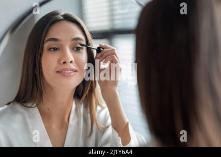 Bella Signora Millennial che fa il trucco quotidiano vicino a specchio in bagno, applicazione Mascara Foto Stock
