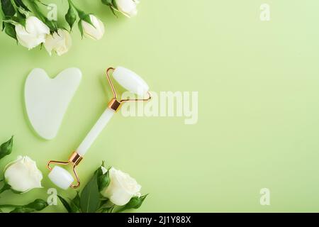 Strumento di massaggio Guasha bianco o pietra da massaggio gua sha e rullo facciale con fiori di rosa su sfondo verde chiaro. Cura della pelle a casa, anti-invecchiamento e sollevamento Foto Stock