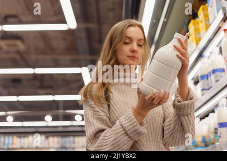 Negozi di alimentari. Donna leggere bottiglia di latte in negozio in piedi vicino scaffale con prodotti lattiero-caseari in supermercato. Indispensabili per l'acquisto di Lady Buyer Foto Stock