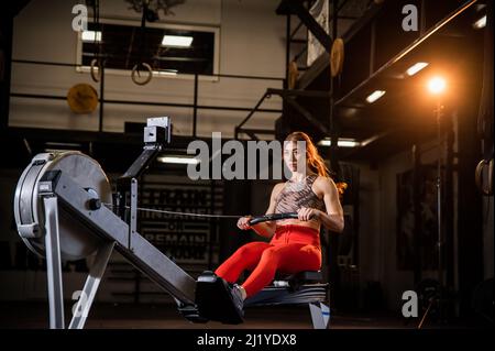 Una donna muscolare attraente in una palestra completamente attrezzata fa allenamento individuale per tutto il corpo su vogatore indoor Foto Stock