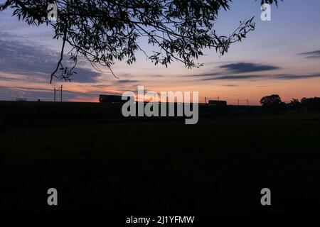 Silhouette di una locomotiva diesel GB Railnight classe 66 che trasporta un treno merci corto sulla linea principale elettrificata della costa occidentale al tramonto. Foto Stock