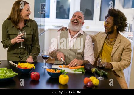 Felice gruppo di amici multigenerazionali divertirsi durante la preparazione di una cena vegetariana, bere vino rosso, concentrarsi sul hipster barbuto uomo Foto Stock