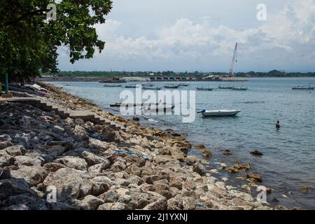 Vista del Porto di Sanur con diverse barche in acqua, una gru sullo sfondo e un uomo che si bagna nel mare. Foto Stock