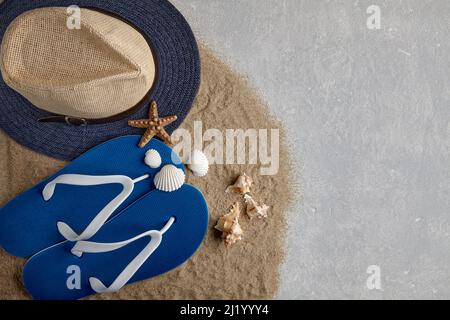 Cappello di paglia, infradito e conchiglie su sabbia di mare sparsa su sfondo di cemento grigio chiaro Foto Stock
