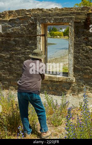 Fotografo che guarda attraverso la finestra di edificio derelitto al Lago Dunstan e Old Cromwell Town, Cromwell, Central Otago, South Island, Nuova Zelanda Foto Stock