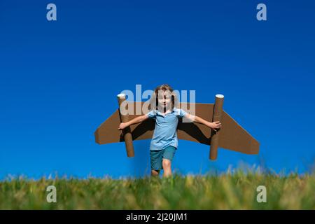 Capretto con super eroe jet pack. Pilota bambino contro sfondo cielo estivo. Ragazzo con volo in aereo di carta, aeroplano giocattolo con ali di cartone, immaginazione Foto Stock