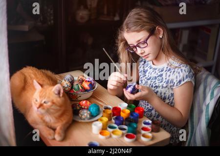 Teen girl dipinge le uova di Pasqua, il suo gatto zenzero giace sul tavolo. Casa interna, Pasqua. Foto Stock