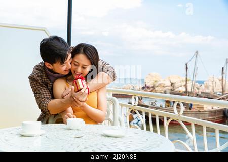 Fidanzata che dà a fidanzato un regalo di fidanzamento a sorpresa in un ristorante vicino a un molo di barche da pesca - foto di scorta Foto Stock