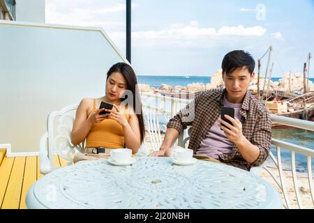 Giovane coppia che utilizza telefoni cellulari al caffè costiero presso il molo di pesca Barca - foto di scorta Foto Stock