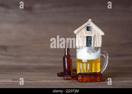 Modello di casa in legno in miniatura in cima a una piccola tazza di birra con bottiglie vuote che si trovano intorno. Abuso di alcool e concetto di homelessness. Foto Stock
