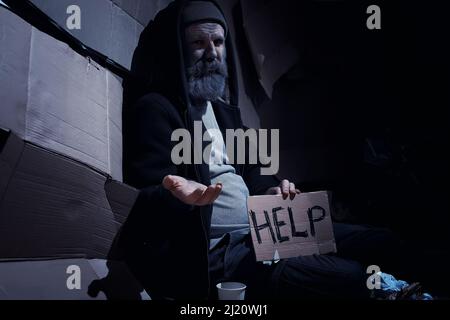 Un uomo senza tetto si siede sulle scatole per strada e chiede aiuto. Ha bisogno di una persona senza tetto chiede soldi per il cibo e durante la notte. Foto Stock