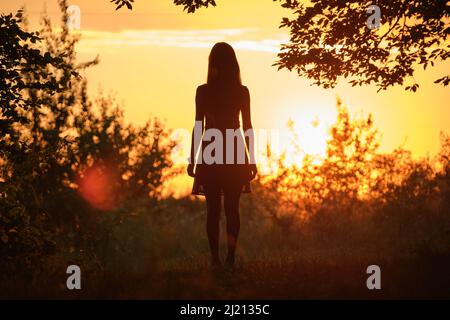 Vista posteriore della giovane donna in abito estivo che cammina da sola attraverso la foresta scura sera. Foto Stock