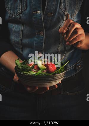 Pranzo vegano sano. Donna in denim blu tiene una ciotola in ceramica con avocado, rucola, pompelmo e insalata di anacardi nelle sue mani. Comfort, pulito, vegetaria Foto Stock