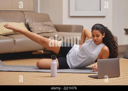 Questo è più difficile di quanto sembri. Scatto completo di una giovane donna che usa il suo laptop per seguire una lezione di fitness online nel suo salotto. Foto Stock