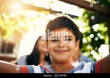 HES ottenere buono a prendere selfie. Ritratto di un adorabile ragazzino che prende un selfie con i genitori sullo sfondo. Foto Stock