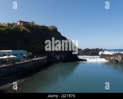 I visitatori si siedono al sole al bar sul lungomare di Seixal Madeira Portogallo UE con le onde dell'Oceano Atlantico che si infrangono sulle rocce vulcaniche Foto Stock