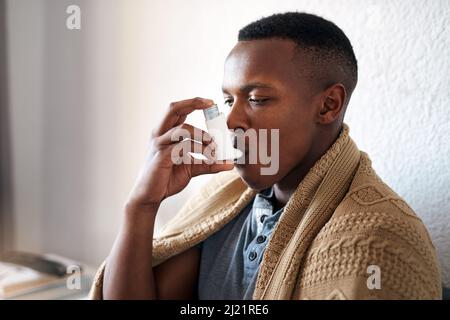 Ho davvero bisogno della mia pompa per l'asma. Scatto corto di un bel giovane uomo seduto da solo nella sua camera da letto e utilizzando una pompa per l'asma. Foto Stock