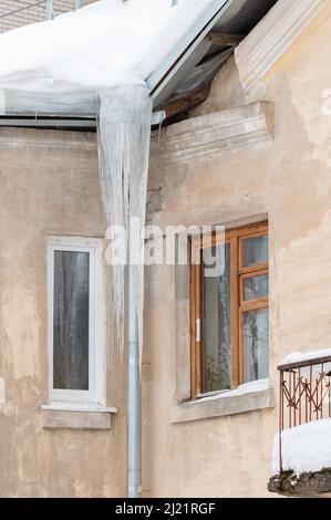 Favolose ghiacciole appendono da un tubo di drenaggio sul bordo del tetto. Sullo sfondo del muro di una vecchia casa di mattoni. Grandi cascate, anche belle file. Nuvoloso giorno d'inverno, luce tenue. Foto Stock