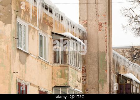 Grandi ghiaccioli appendono dal bordo del tetto. Sullo sfondo del muro di una vecchia casa di mattoni. Grandi cascate, anche belle file. Nuvoloso giorno d'inverno, luce tenue. Foto Stock