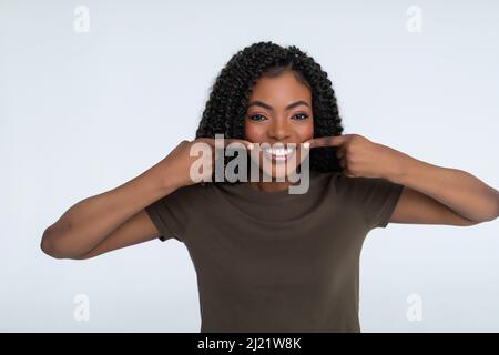 Clinica dentale e concetto di persone. Sorridente donna africana puntando le dita al sorriso, denti bianchi, in piedi in t-shirt su sfondo bianco Foto Stock