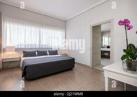 Camera da letto con un grande letto matrimoniale con cuscini grigi per abbinarsi al copriletto, una scrivania di legno bianco, lampade doppie sui comodini e una grande finestra ab Foto Stock