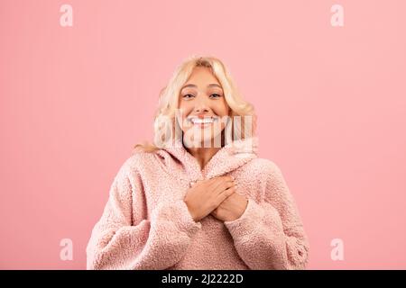 Signora grata che tiene le mani sul petto, esprimendo gratitudine, posando su sfondo rosa studio e sorridendo alla macchina fotografica Foto Stock