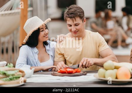Giovani coppie diverse che cucinano insieme vicino camper van all'aperto, tagliando verdure per la prima colazione, godendo viaggio in campeggio Foto Stock