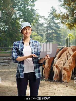 Moderna agricoltore femminile con un computer portatile sorridente e posato su una fattoria di stoppini con cavalli nella parte posteriore Foto Stock