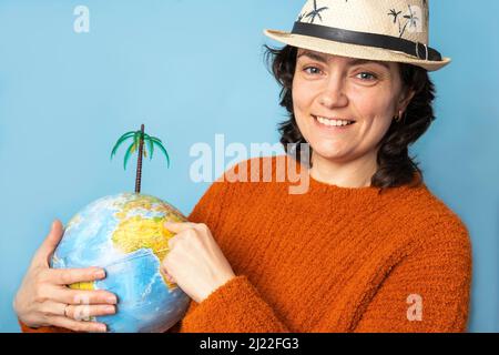 Una donna graziosa in un cappello di sole tiene un globo nelle sue mani e mostra un luogo di vacanza Foto Stock