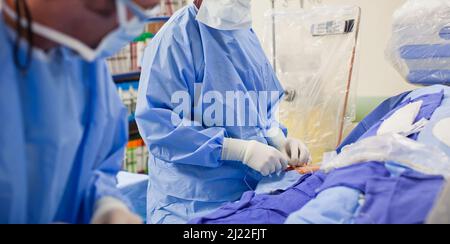 Due medici chirurgo non descritti che fanno un intervento chirurgico in una stanza chirurgica indossando scrub blu e guanti in lattice bianco Foto Stock