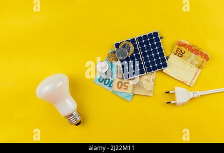 Pannello fotovoltaico, denaro e filo di spina isolati su sfondo giallo. Pannello olar con vari valori di denaro brasiliano. Bus energia pulita alternativo Foto Stock