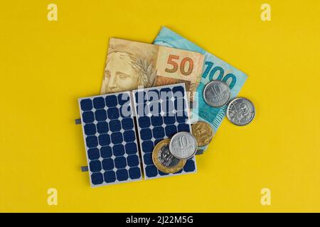 Pannello fotovoltaico, denaro e filo di spina isolati su sfondo giallo. Pannello olar con vari valori di denaro brasiliano. Bus energia pulita alternativo Foto Stock