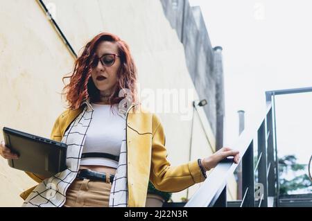 Intraprendente, riuscita e potenziata giovane donna rossa pronta ad andare al lavoro, scendendo le scale con il cielo sullo sfondo, in giallo casual Foto Stock