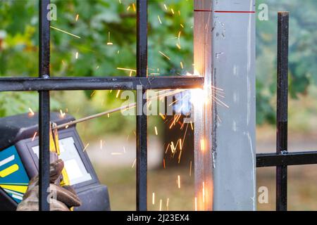Scintille brillanti dall'arco di saldatura elettrica volano in direzioni diverse contro lo sfondo di una recinzione metallica e di un saldatore in funzione in sfocatura. Foto Stock