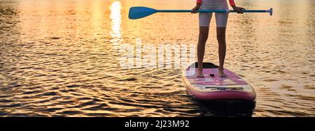 Primo piano di una giovane donna con gambe sottili in piedi su tavola sup con paddle lunghe in mani. Donna attiva in abbigliamento sportivo che passa la sera per praticare sport acquatici. Foto Stock