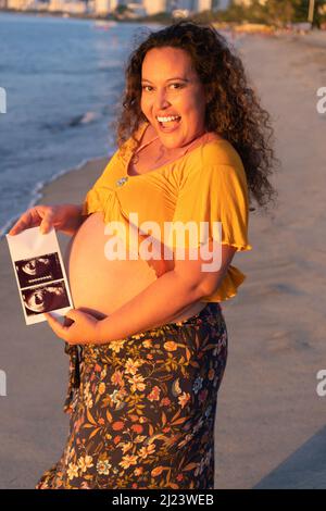 Donna incinta in piedi e tenendo una scansione ecografica sul ventre. Concetto di gravidanza, paternità e preparazione Foto Stock