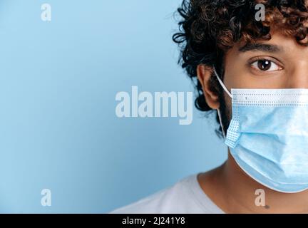 Prevenzione delle infezioni. Mezzo volto di un giovane ragazzo curly indiano o arabo adulto, con una maschera medica protettiva sul suo volto, guardando la fotocamera mentre si trova su un isolato sfondo blu Foto Stock