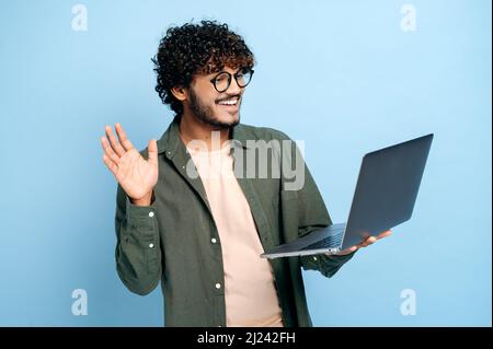 Comunicazione video online. Il tipo indiano o arabo, in abiti casual, freelancer o studente, utilizza un computer portatile per una videoconferenza, si erge su sfondo blu isolato, saluta un interlocutore, sorridendo Foto Stock