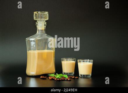 Liquore di caffè cremoso, alcol forte in una bottiglia e due bicchieri su sfondo nero, accanto alle foglie di menta e chicchi di caffè. Primo piano. Foto Stock