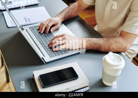 Le mani di un giovane uomo d'affari che digita sulla tastiera del laptop sul posto di lavoro, analizzando al contempo i dati finanziari online Foto Stock