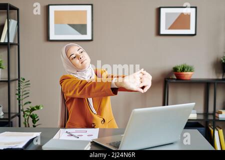 Giovane lavoratrice in ufficio stanca che tiene le braccia tese di fronte a se stessa mentre fa l'esercizio davanti al computer portatile dalla scrivania Foto Stock