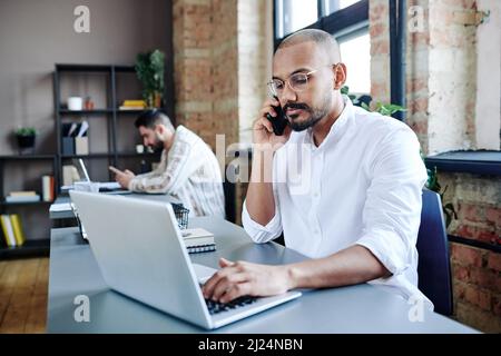 Giovane impiegato impegnato in casualawear che parla al telefono cellulare dal posto di lavoro mentre si siede di fronte al computer portatile e digitando Foto Stock