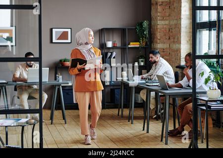 Giovane donna d'affari contemporanea in hijab camminando lungo le scrivanie di colleghi maschi che lavorano davanti ai computer in un ufficio moderno Foto Stock