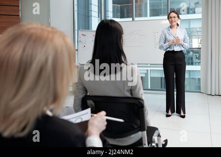 Giovane vettura sorridente di affari in formalwear che comunica con il pubblico al seminario mentre si levano in piedi dalla lavagna bianca con i grafici Foto Stock