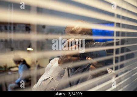 Giovane agente afro-americano FBI o detective con fotocamera che scatta foto attraverso le persiane veneziane mentre si trova accanto alla finestra dell'ufficio Foto Stock