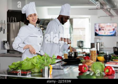 Cuochi professionali che indossano uniformi di cottura per preparare verdure fresche guarnire per un delizioso piatto da pranzo. Lavoratori dell'industria alimentare multirazziale in piedi nella cucina professionale del ristorante. Concetto di cucina Foto Stock