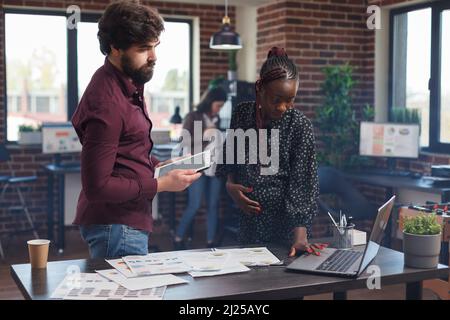Dipendenti aziendali multietnici che cercano di risolvere problemi finanziari utilizzando tablet e notebook. Gravidanza madre che lavora sul computer moderno mentre analizzano le idee di strategia di marketing. Foto Stock